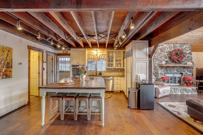 Open-concept rustic cabin kitchen and living room with exposed wood ceiling beams, large island with stools, farmhouse cabinets, and a stone fireplace decorated with a red wreath.