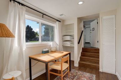 Cozy sunlit home office nook by a large window with a wooden desk and chair, table lamp, patterned rug, built-in shelves with linens, and a short stairway to an upper entry.