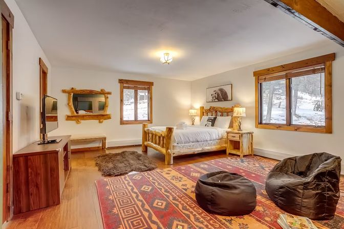 Cozy rustic mountain cabin bedroom with log bed and nightstands, patterned Southwestern rug, bean bag chairs, TV stand and large wood-trim windows framing a snowy forest view