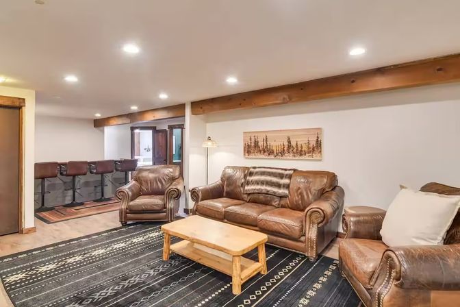 Cozy rustic basement living room with brown leather sofa and armchairs, wooden coffee table on a dark patterned rug, bar counter with stools, exposed wood beams and recessed lighting.