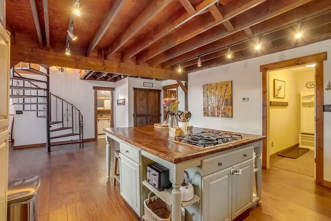 Cozy rustic kitchen with exposed wooden beams, hardwood floors, large butcher-block island with gas cooktop and pale painted base, track lighting, and a black spiral staircase by the pantry door.