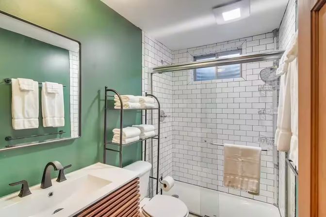 Clean, spa-like modern bathroom with white subway-tile shower and glass sliding door, green accent wall, black-framed mirror and matte-black fixtures, wooden-slat vanity, metal shelving stacked with folded white towels and a toilet.