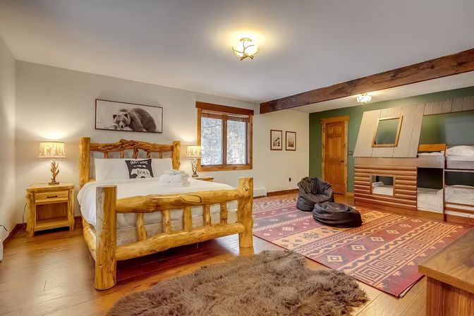 Cozy mountain cabin bedroom with handcrafted log king bed, built-in bunk beds with cabin-style paneling, patterned southwestern rug, beanbag chairs, rustic wood beam and bear wall art