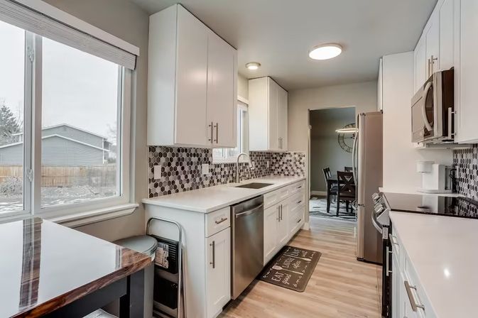 Bright modern galley kitchen in a suburban home with white cabinets, hexagon black-and-gray tile backsplash, stainless appliances, sink under a window and wood-look floors