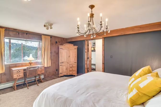 Cozy rustic bedroom with wood-paneled walls, white bed topped with yellow accent pillows, antique wooden armoire and desk by a window overlooking a mountain view, and a crystal chandelier overhead.