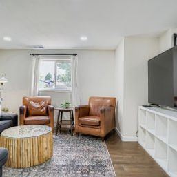 Sunlit modern living room with two brown leather armchairs, round wooden coffee table, patterned area rug, flat-screen TV on white cube shelving, and hardwood floors.