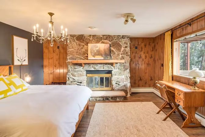 Cozy rustic mountain-cabin bedroom with a stone fireplace, wood-paneled walls, white bed with yellow accent pillows, chandelier and a wooden desk by a window overlooking a pine forest.