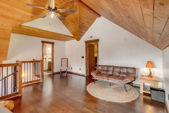 Cozy rustic loft living area with sloped wood-paneled vaulted ceiling, hardwood floors, ceiling fan, leather sofa on a round jute rug, wooden side table with lamp, and open railing overlooking stairs.
