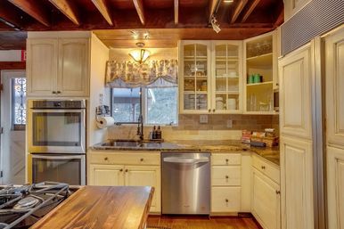 Cozy country-style kitchen with exposed wood beam ceiling, cream cabinets, granite countertops, tile backsplash, stainless double oven and dishwasher, farmhouse sink beneath a patterned valance, and a wooden island in the foreground.