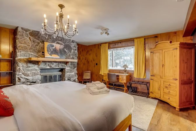 Cozy rustic cabin bedroom with stone fireplace and wood-paneled walls, chandelier-lit king bed with white linens and folded towels, yellow-curtained window overlooking snowy trees, wooden desk and armoire.