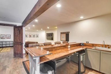 Cozy finished basement home bar with polished wood countertop, leather bar stools, mini fridges, sink, Scrabble wall board, recessed lighting and open lounge area