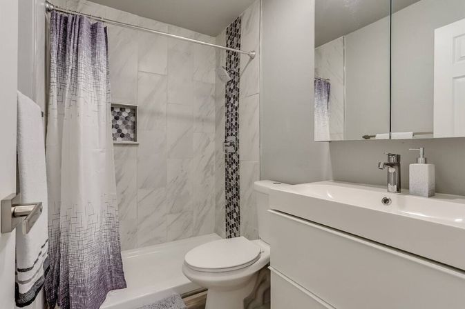 Crisp small modern bathroom with white marble-tiled shower, vertical hexagon mosaic accent and niche, purple-ombre shower curtain, white tub and toilet, and sleek white vanity with integrated sink and mirrored cabinet.