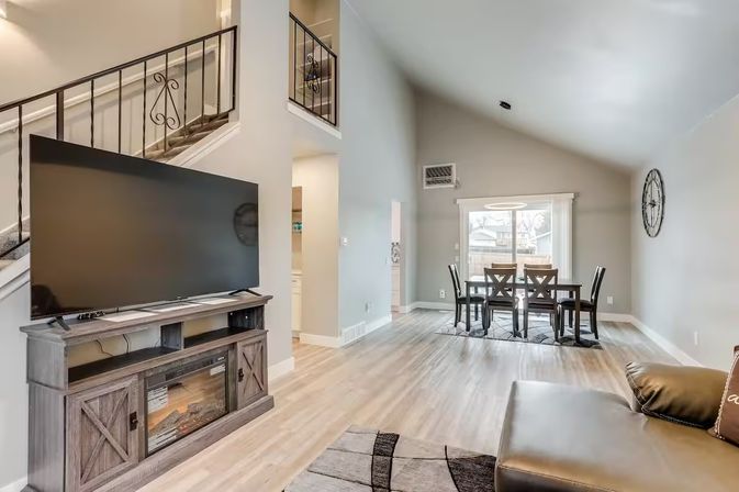 Open-concept living and dining area in a modern suburban home with vaulted ceiling, light wood floors, large flat-screen TV on a rustic media console with electric fireplace, wrought-iron staircase railing, and a six-seat dining table by sliding glass doors.