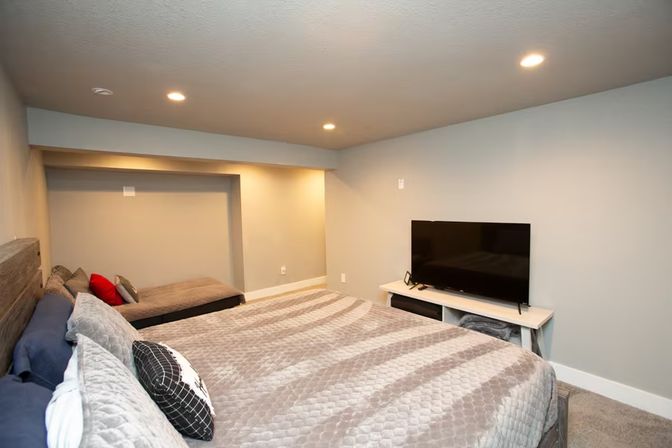 Cozy modern finished basement bedroom with low ceiling, neutral gray walls, recessed lighting, large quilted gray bed with accent pillows, small daybed in an alcove with a red throw pillow, and a flat-screen TV on a white console.