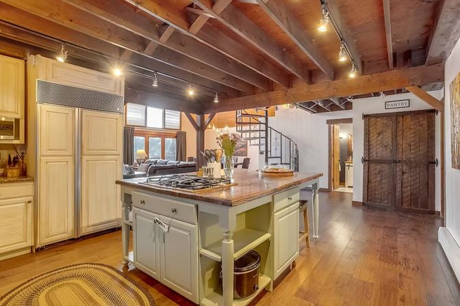 Rustic open-concept kitchen with exposed wood beams, farmhouse island with gas cooktop, hardwood floors, spiral staircase, sliding barn-door pantry, and natural light
