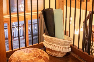 Cozy rustic loft scene with a tasseled woven basket holding rolled black and mint yoga mats on a wooden railing, warm hanging Edison lights and a leather pouf nearby.