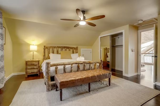 Cozy rustic cabin-style loft bedroom with a log bed, tufted leather bench, bedside lamp, ceiling fan, mirrored closet and balcony access
