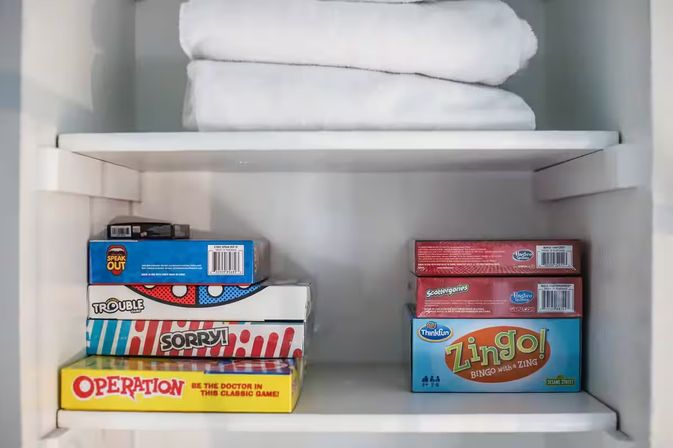 Linen closet shelf with folded white towels above a neat stack of colorful family board games, including Operation, Sorry, Trouble, Speak Out, Scattergories and Zingo.