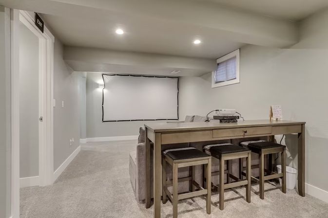Cozy finished basement home theater with large projector screen, projector on a console table, three bar stools, gray sofa, carpeted floor and recessed ceiling lights.