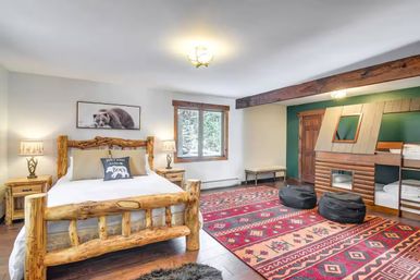 Cozy mountain cabin bedroom with rustic log bed, bedside lamps, forest-view window, colorful Southwestern rugs, built-in bunk beds, beanbags and playful bear-themed wall art.