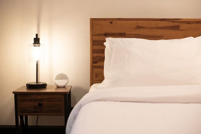 Cozy modern bedroom interior with white linens, a wooden headboard, and a bedside table holding a glowing glass lamp and round alarm clock.