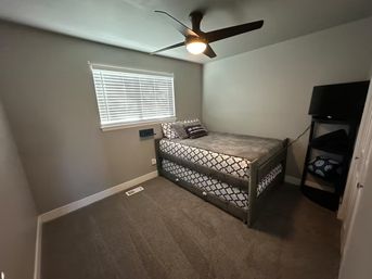 Cozy small bedroom with gray walls and carpet, twin trundle bed with black-and-white patterned bedding, modern black ceiling fan with light, white window blinds and a corner shelf with TV and linens.