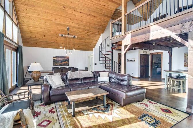 Sunlit rustic mountain-cabin living room with vaulted wood ceiling, brown leather sectional, patterned rug and spiral staircase to an open loft