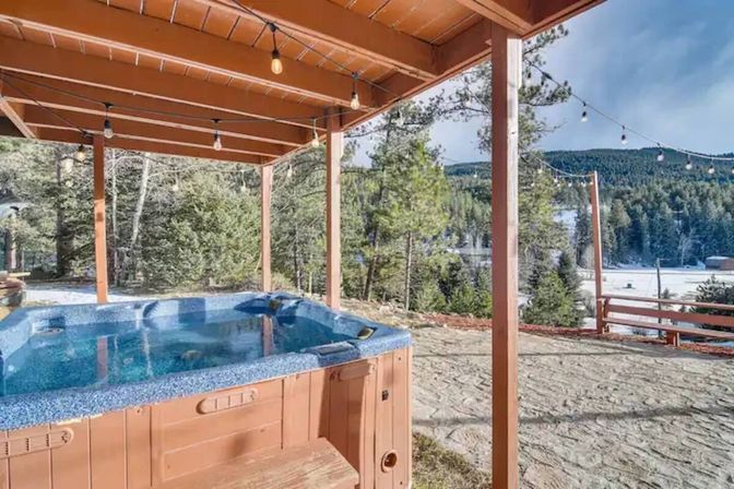 Outdoor hot tub under a wooden pergola with string lights, overlooking pine forest and a snowy mountain lake