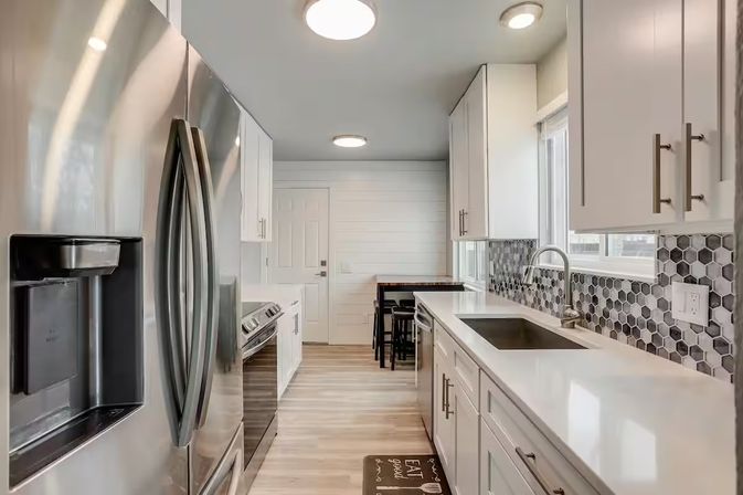 Bright modern galley kitchen with stainless steel French-door refrigerator, white cabinets, hexagonal gray tile backsplash, quartz countertops, undermount sink and a small breakfast nook with stools