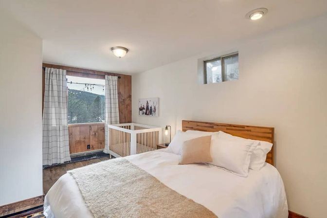 Cozy mountain-cabin bedroom with wooden headboard, white bedding and beige throw, small white crib by a window with string lights and a forested mountain view, neutral curtains and warm wood accents.