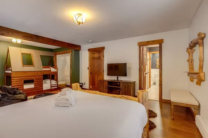 Cozy rustic mountain-lodge bedroom with log-frame king bed, cabin-style bunk beds, flat-screen TV, wood floors and trim, and an en-suite bathroom — family-friendly vacation rental interior.