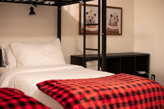 Cozy bedroom interior with a lower bunk bed in crisp white linens, red-and-black buffalo plaid throws, black metal ladder and cubby shelving, and framed bear prints on a neutral wall.