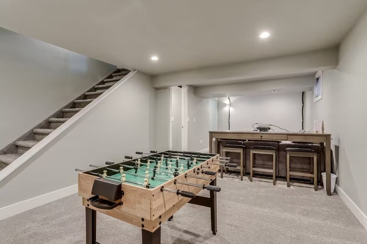 Finished residential basement rec room with a foosball table in the foreground, carpeted stairs to the left, neutral gray walls with recessed lighting, and a wooden bar with three stools.