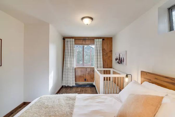 Cozy rustic bedroom-nursery with a white bed and wooden headboard, small white crib by a wood-paneled window with check curtains, hardwood floor and soft neutral accents.
