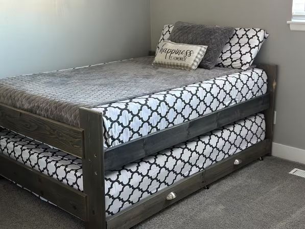 Gray wooden trundle bed with black-and-white trellis-pattern mattresses, gray quilted comforter and 'Happiness is homemade' accent pillow in a neutral gray carpeted bedroom
