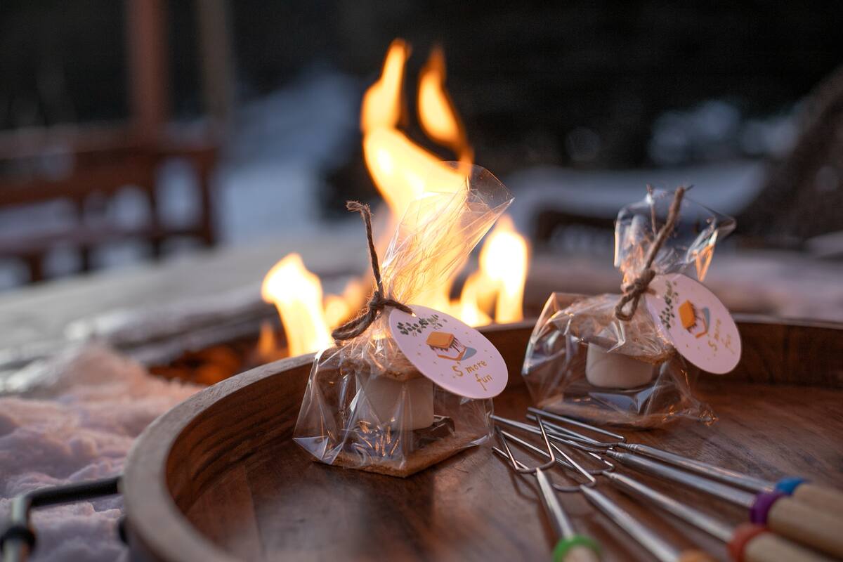 Cozy winter scene: two clear-wrapped s'mores kits tied with twine on a wooden tray, colorful marshmallow roasting forks nearby, glowing outdoor fire pit and snow in the background.