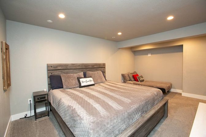 Cozy finished basement bedroom with king-size wooden bed, gray quilted bedding, recessed ceiling lights and an alcove daybed with a red accent pillow