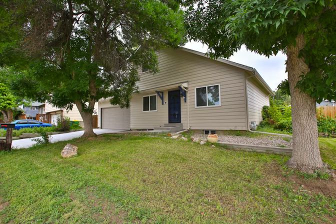 Beige single-story suburban house with two-car garage, blue front door, grassy front yard, rock landscaping and mature trees along the driveway in a residential neighborhood.