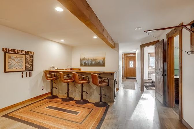 Cozy rustic home bar with four leather swivel stools at a wooden counter, Scrabble wall display, exposed beam and runner rug leading down a hardwood hallway to a wooden door.