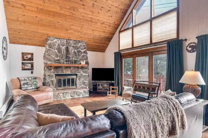 Cozy mountain cabin living room with rustic stone fireplace, vaulted wood ceiling, leather sectional with fur throw, wooden coffee table and large windows opening to a forested deck.