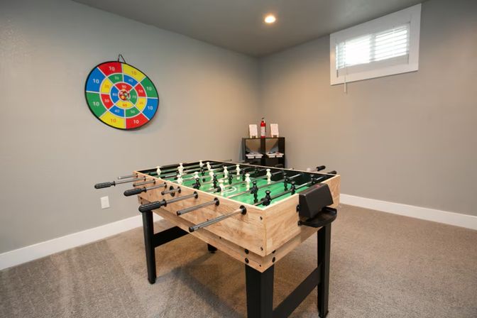 Basement game room with wooden foosball table (green field), colorful circular dartboard on a gray wall, carpeted floor and a small high window.