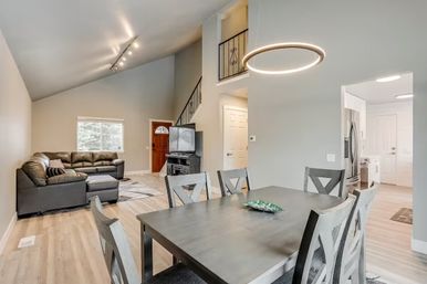 Open-concept living and dining area in a modern home with vaulted ceiling, circular pendant light, hardwood floors, gray leather sectional, dining table and visible kitchen doorway.