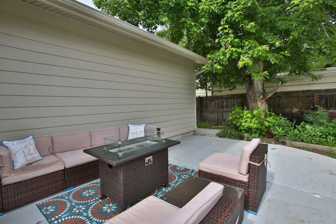 Cozy backyard concrete patio with wicker sectional and chairs in blush cushions arranged around a glass-top fire table on a colorful outdoor rug, shaded by a large tree and bordered by a wooden privacy fence and garden beds.