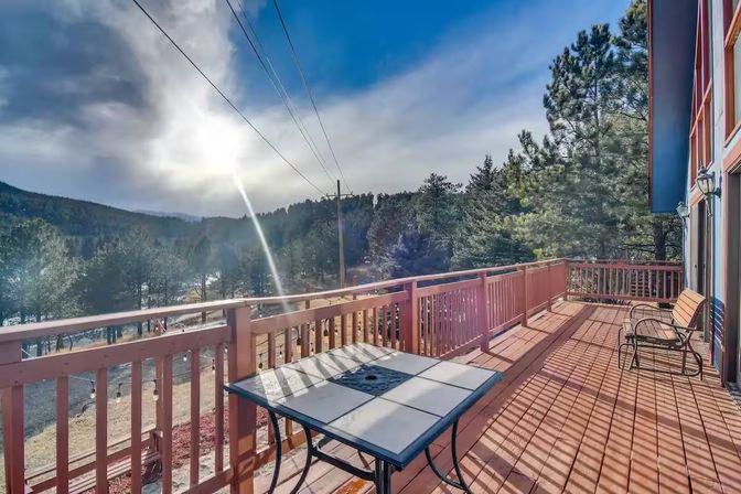 Cozy sun-drenched wooden deck with a tiled patio table and metal chairs, red railing overlooking pine-covered foothills and distant mountains under a bright blue sky with sun glare.