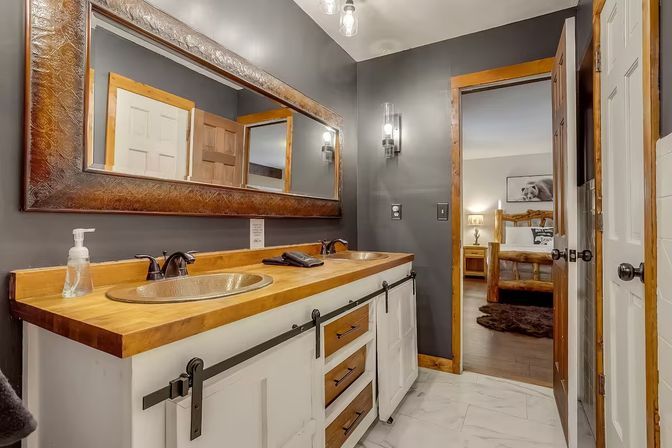 Rustic double-vanity bathroom with wood countertop and copper sinks, black faucets, sliding barn-style cabinet hardware, large ornate mirror, dark gray walls, marble-look floor tile and an open door showing a log-bed bedroom.