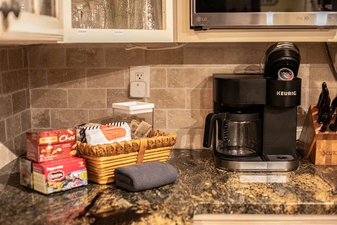 Cozy home kitchen coffee corner on a granite countertop with a black single-serve coffee maker and glass carafe, wicker basket of tea and coffee supplies, boxed tea, knife block and tiled backsplash.