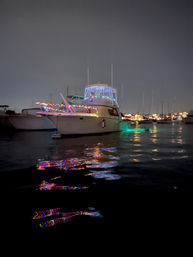 White yacht moored in a marina at night, decorated with colorful string lights and green underwater glow, vibrant multicolor reflections rippling across dark harbor water with sailboats and dock lights in the background.
