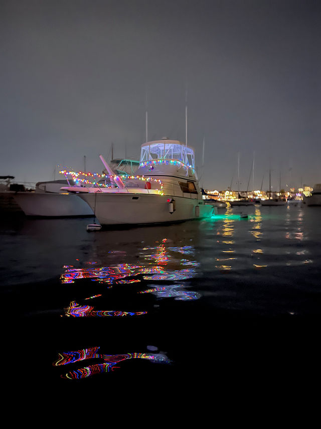 White yacht moored in a marina at night, decorated with colorful string lights and green underwater glow, vibrant multicolor reflections rippling across dark harbor water with sailboats and dock lights in the background.
