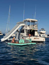 White motor yacht anchored in calm blue ocean with a large gray inflatable water slide off the stern and three people lounging on a mint-green floating platform under a clear sunny sky.