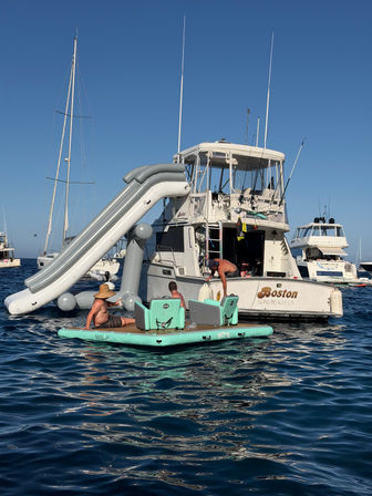 White motor yacht anchored in calm blue ocean with a large gray inflatable water slide off the stern and three people lounging on a mint-green floating platform under a clear sunny sky.